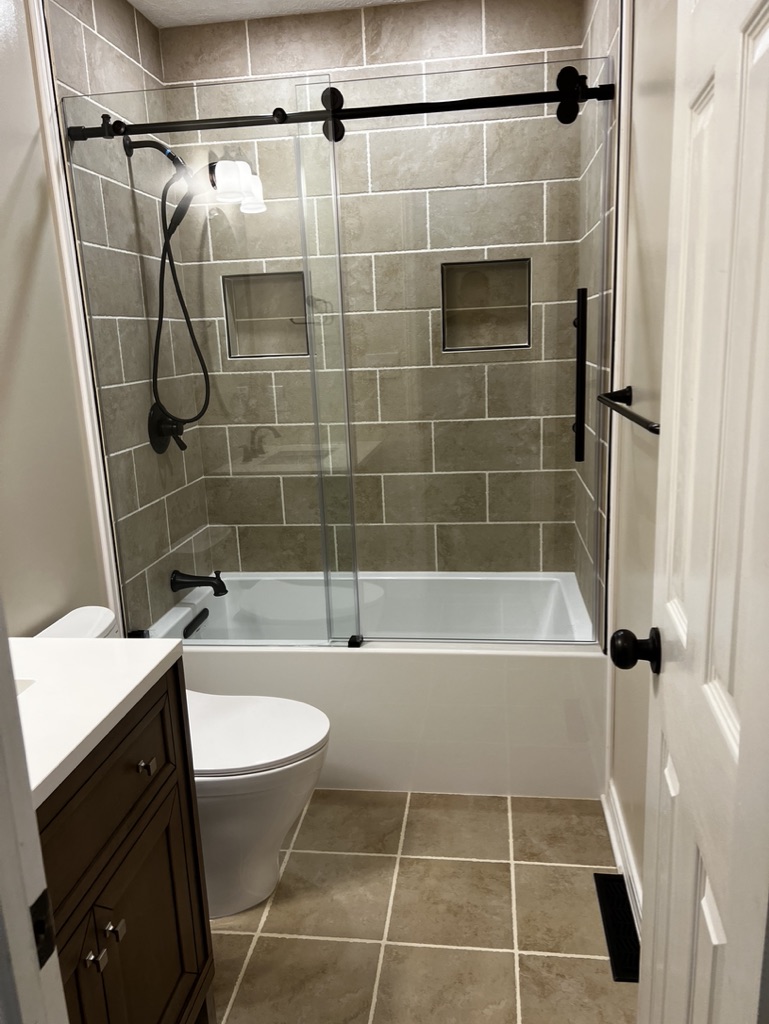 Completed bathroom with tiled tub and shower surround, sliding glass enclosure, recessed niches, vanity, toilet, and tile flooring